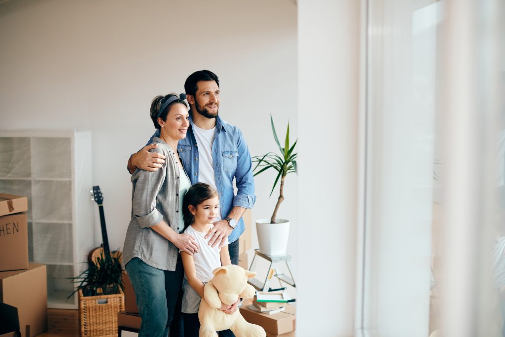 A family of three stands together in a new home surrounded by moving boxes.